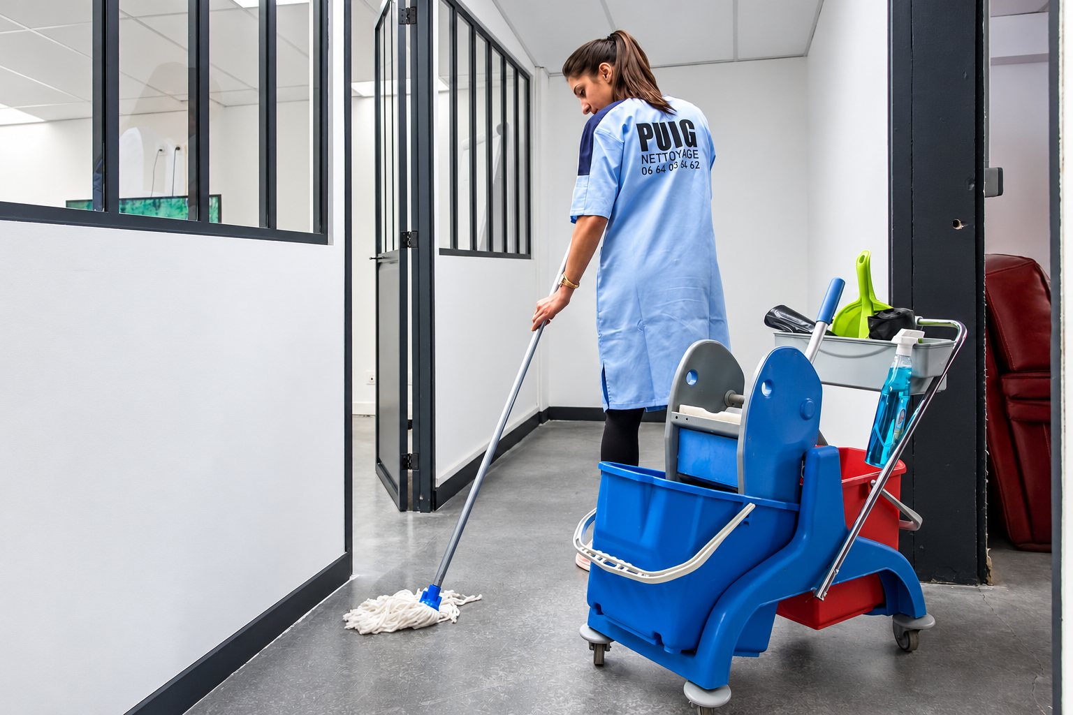 Une agente en blouse bleue passe la serpillière dans un couloir de bureaux avec son chariot de ménage bleu et rouge.