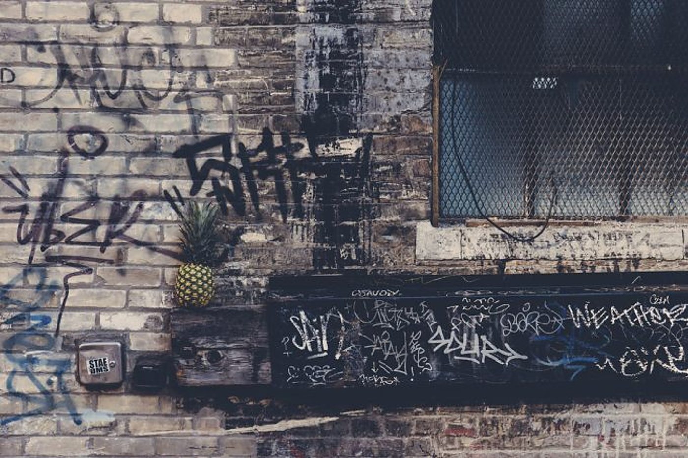 Un ananas est posé sur une poutre en bois devant un vieux mur de briques recouvert de graffitis, à côté d'une fenêtre grillagée.