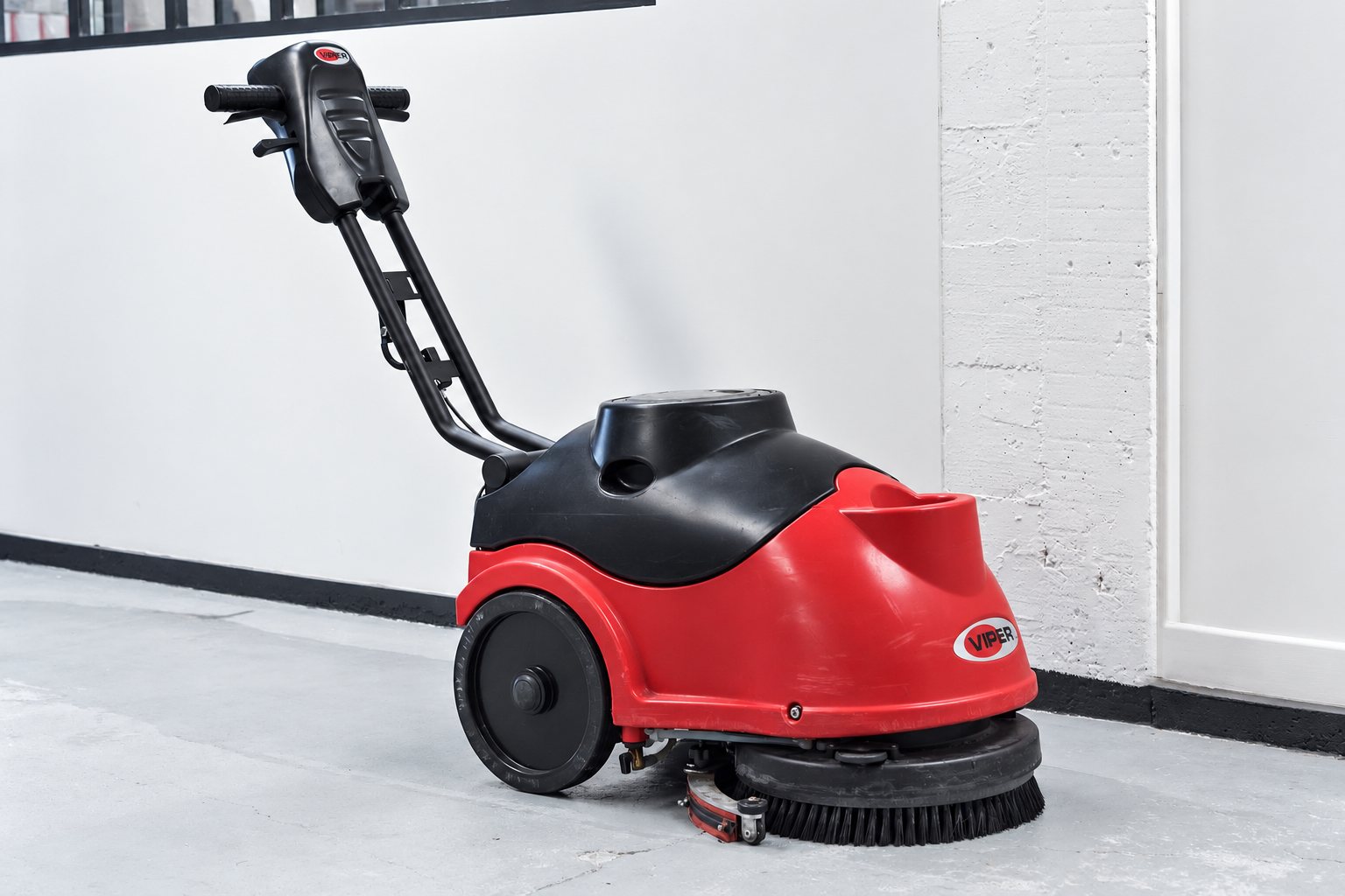 Une autolaveuse compacte rouge et noire de marque Viper, avec sa brosse ronde, est posée sur un sol en béton devant un mur blanc.