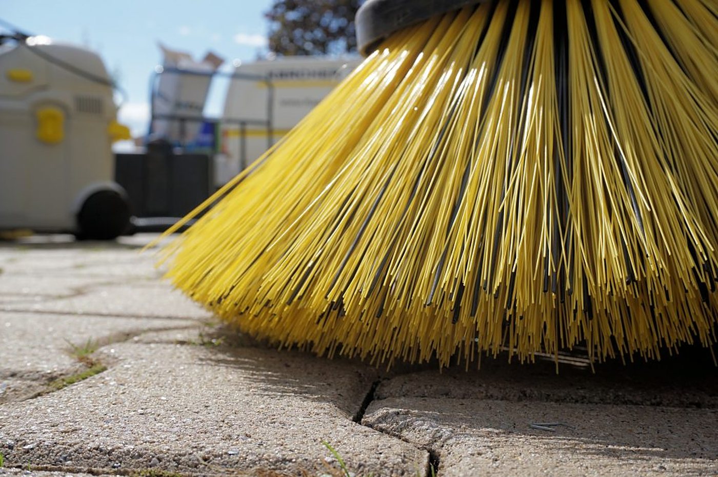 Gros plan sur les poils jaunes d'une brosse de balayeuse posée sur un sol pavé, avec des véhicules de nettoyage flous en arrière-plan.