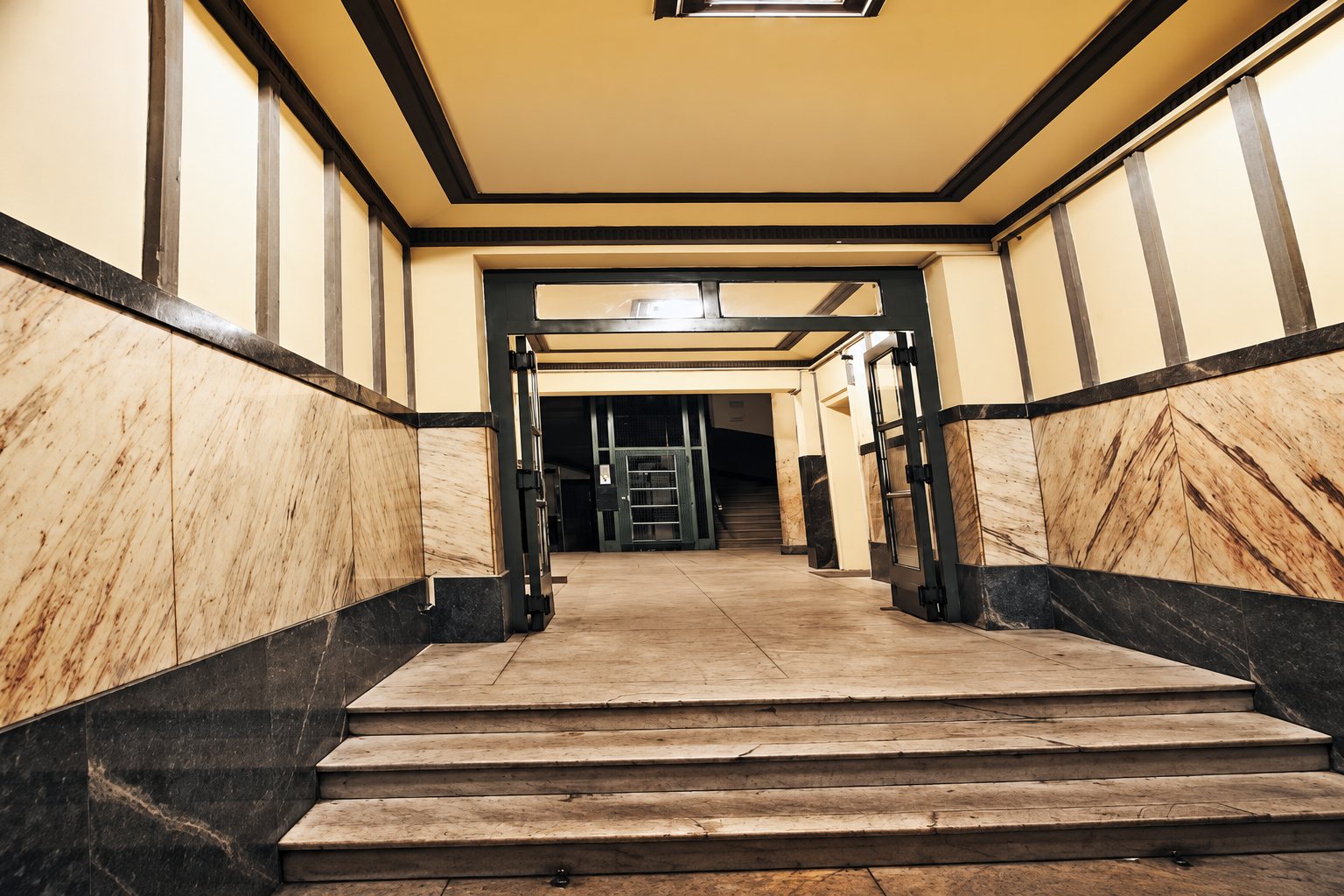 Un couloir d'entrée aux murs de marbre beige et noir, avec quelques marches, des portes vitrées à cadre sombre et un éclairage jaune chaleureux.