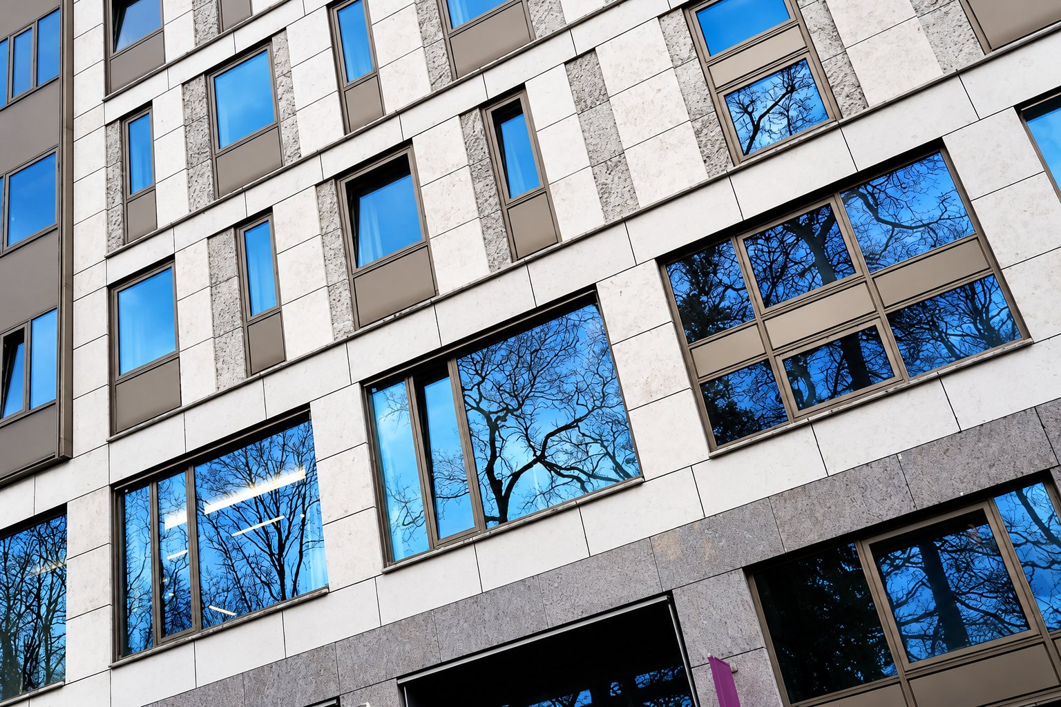 Façade d'un immeuble moderne en pierre claire dont les fenêtres reflètent des arbres et le ciel bleu, vue en contre-plongée.