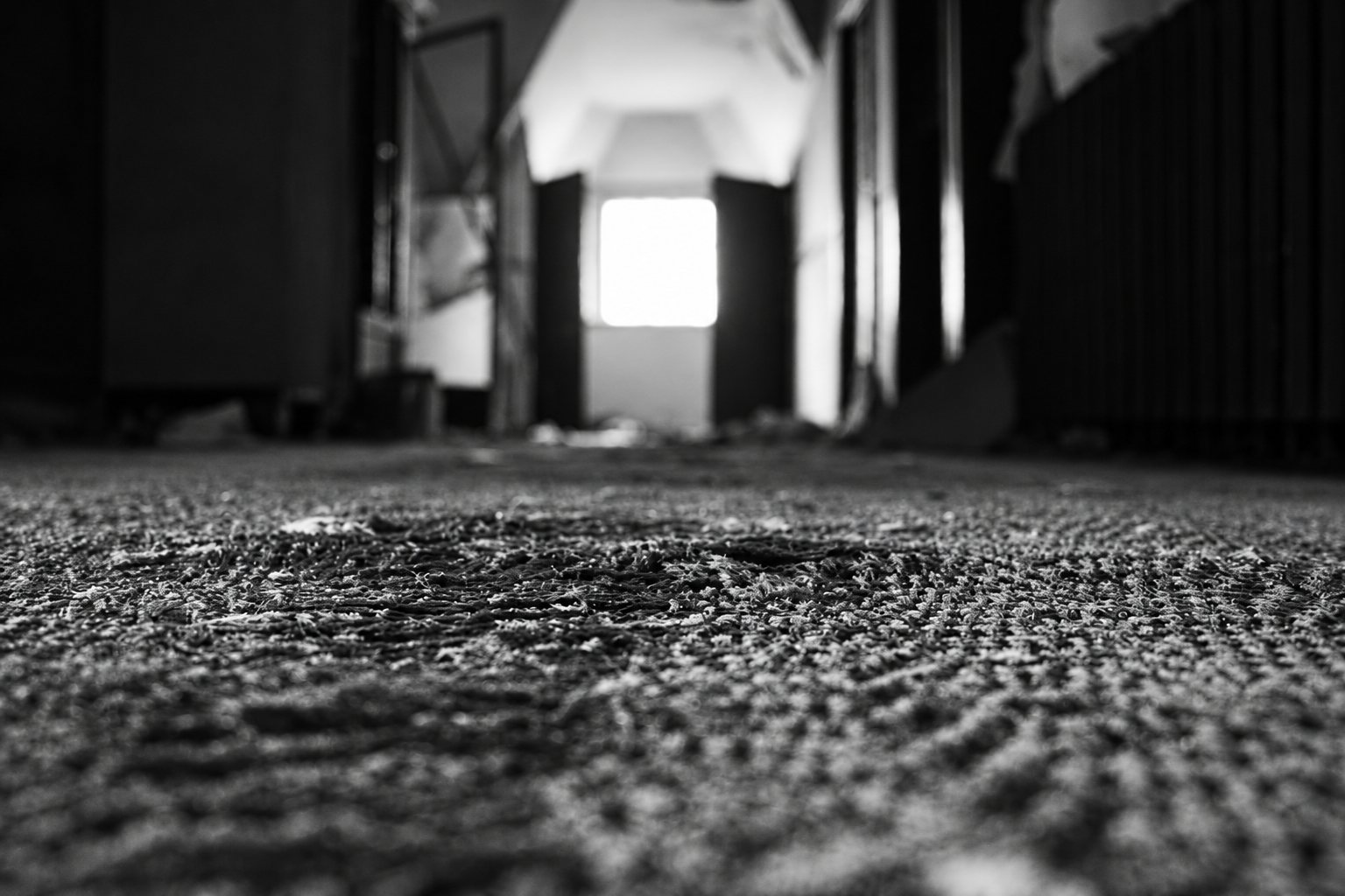 Photo en noir et blanc d'une moquette usée prise au niveau du sol, avec un couloir sombre et une fenêtre lumineuse au fond.