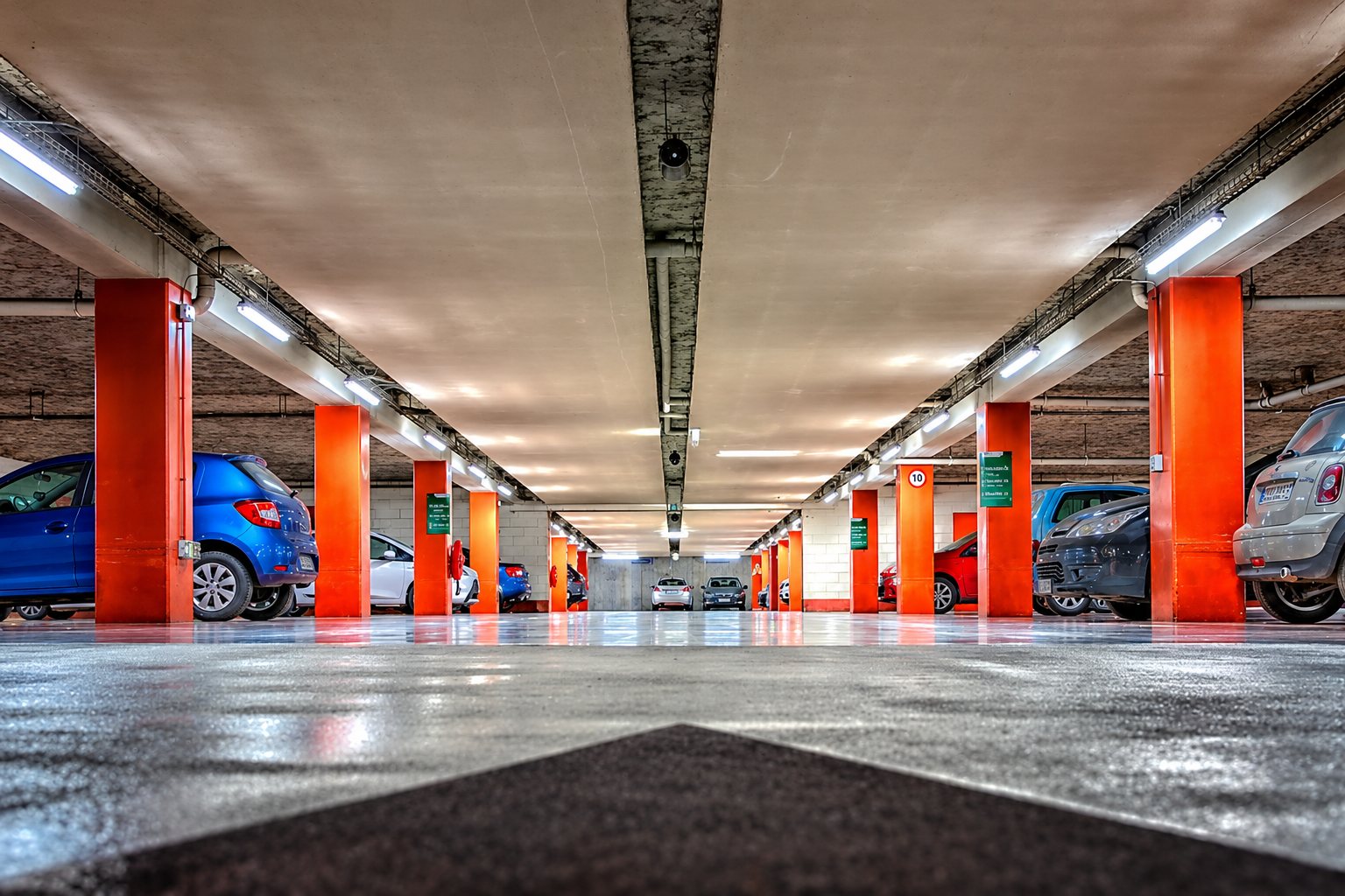 Vue au ras du sol d'un parking souterrain éclairé aux néons, avec des piliers orange et plusieurs voitures stationnées de chaque côté.