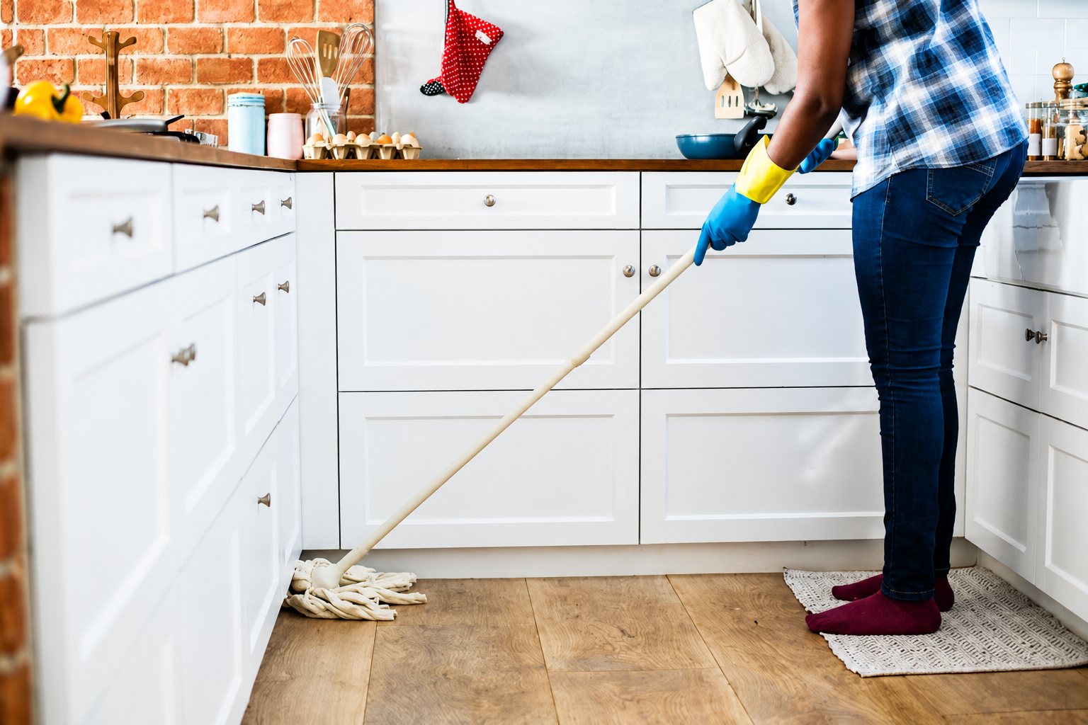 Une personne portant des gants en caoutchouc bleus et jaunes passe la serpillière sur le parquet d'une cuisine aux meubles blancs et au mur de briques.