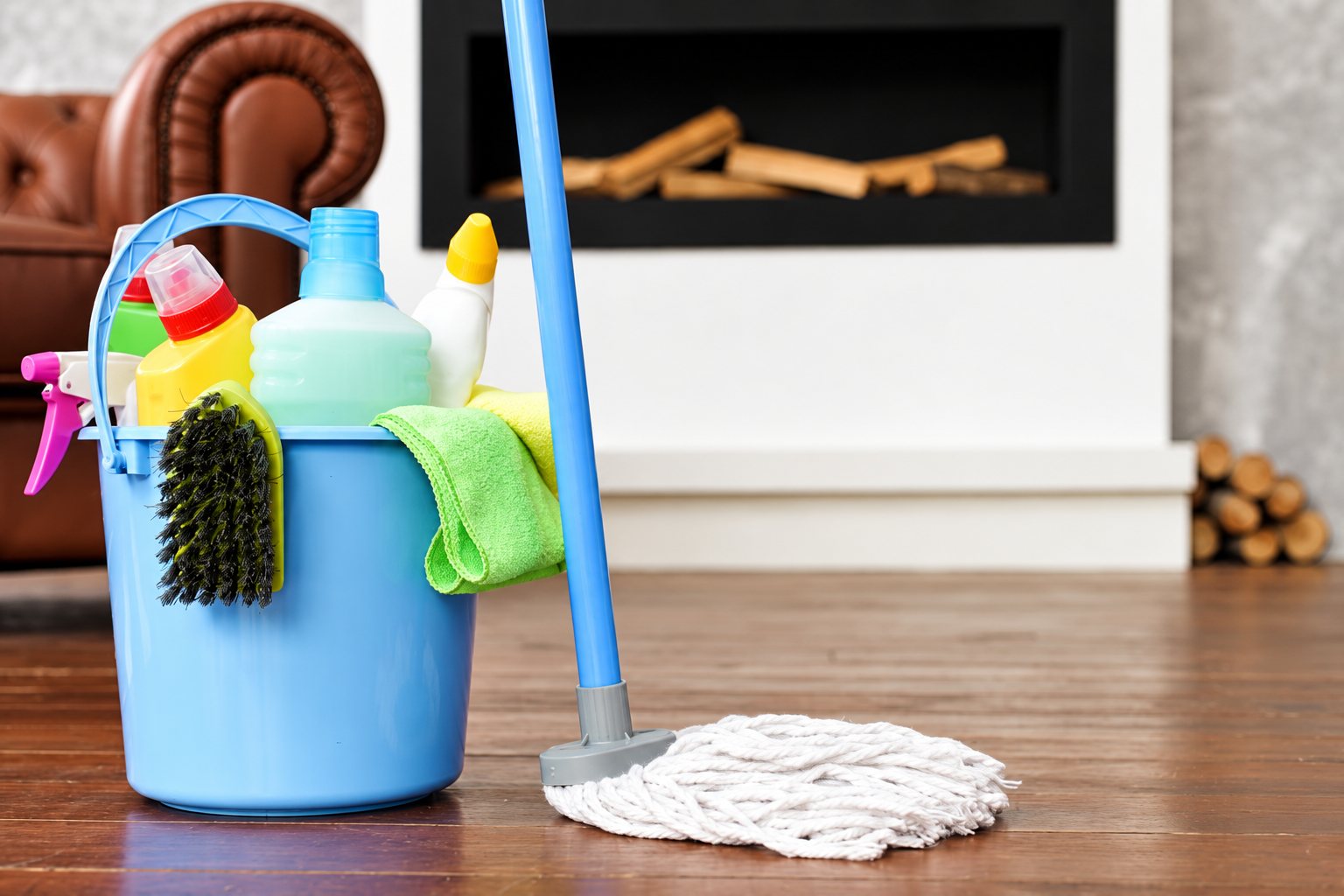 Un seau bleu rempli de flacons de produits ménagers, d'une brosse et d'un chiffon vert, posé à côté d'un balai à franges sur un parquet devant une cheminée.