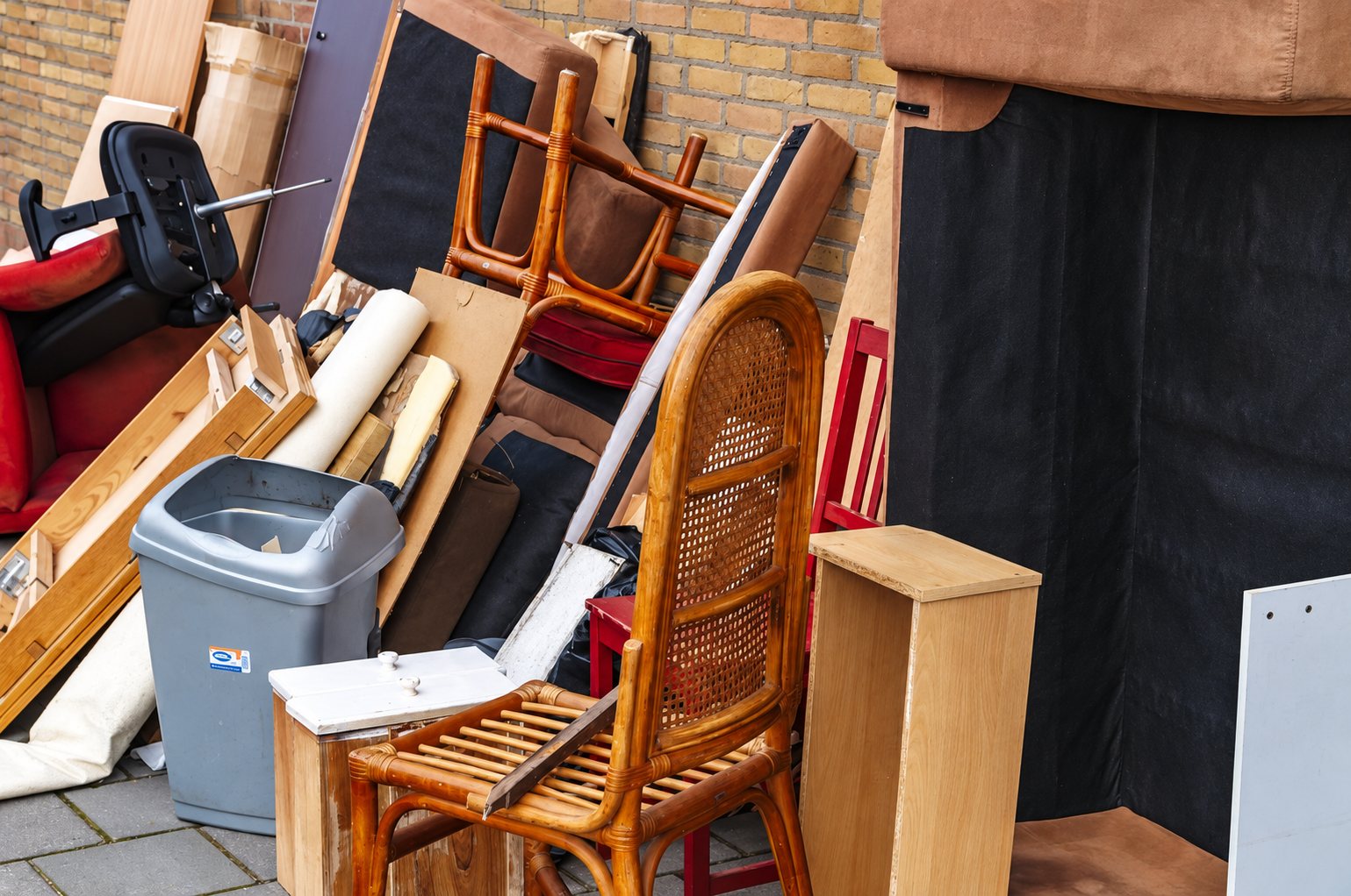 Un tas de vieux meubles, dont une chaise en rotin, une poubelle grise et des panneaux de bois, est entassé sur le trottoir contre un mur de briques.
