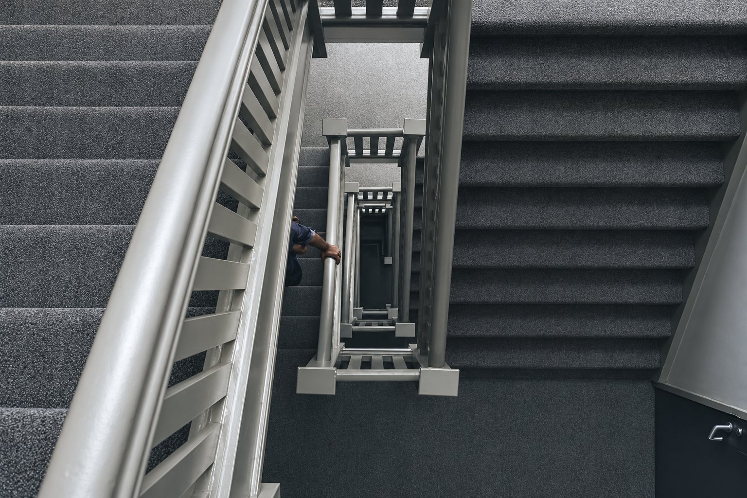 Vue plongeante sur une cage d'escalier recouverte de moquette grise, avec une rampe à barreaux et une personne appuyée plus bas.