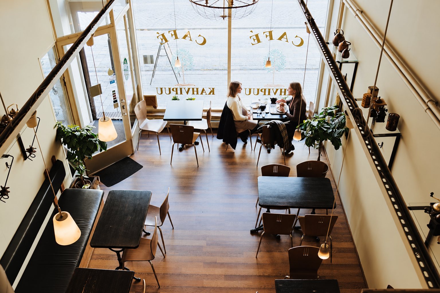 Vue depuis l'étage d'un café aux tables sombres et au parquet, où deux femmes discutent près de la grande vitrine marquée Café Kahvipuu.